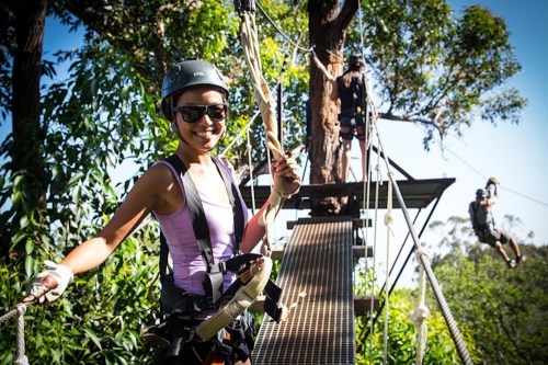 A smiling woman in a helmet and sunglasses stands on a zipline platform, ready to zip through a forested area.