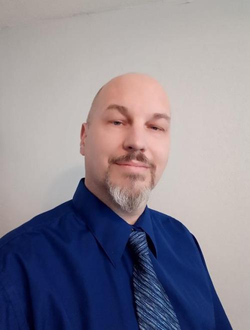 A man with a bald head and goatee smiles, wearing a blue dress shirt and tie against a light background.