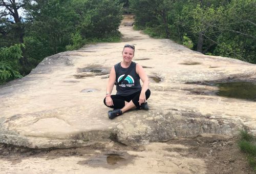 A person sits on a rocky ledge surrounded by greenery, smiling and enjoying the outdoors.