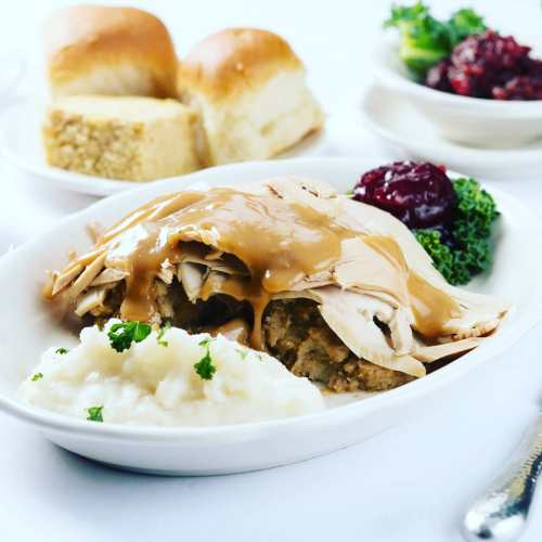 A plate of turkey with gravy over stuffing, mashed potatoes, and garnished with kale, alongside rolls and cranberry sauce.