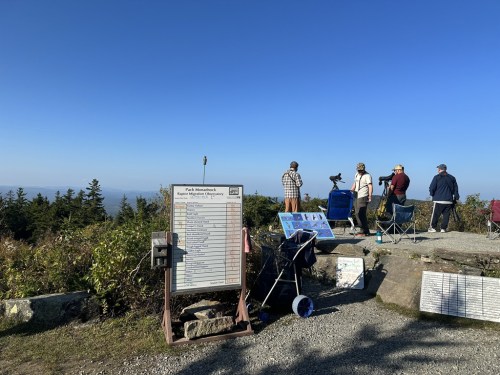 A group of people observing a scenic view from a mountaintop, with informational signs and equipment nearby.