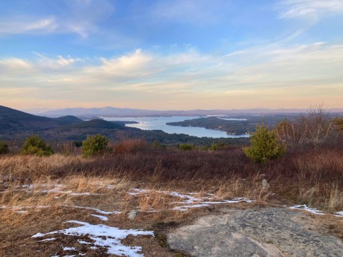 A scenic view of a lake surrounded by hills and trees, with a clear sky and patches of snow on the ground.