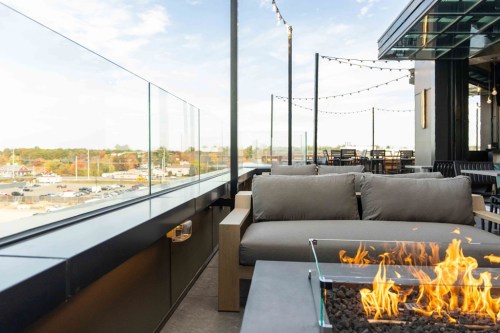 A rooftop lounge with a cozy sofa and a fire pit, overlooking a scenic view and string lights in the background.