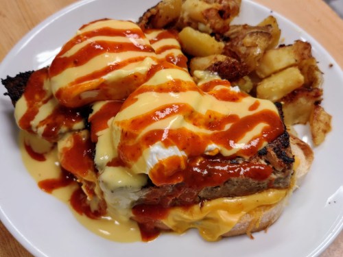 A plate of poached eggs on toast topped with creamy sauce and drizzled with hot sauce, served with crispy potatoes.