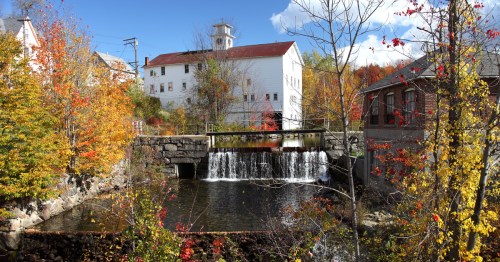 A scenic view of a small waterfall flowing into a pond, surrounded by colorful autumn foliage and historic buildings.