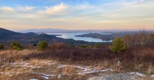 A scenic view of a lake surrounded by mountains and trees, with a soft sunset sky in the background.