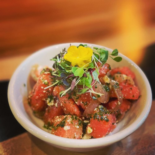 A bowl of watermelon salad topped with herbs, microgreens, and a yellow flower, served in a rustic dish.
