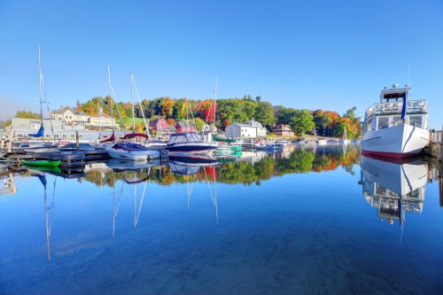 A serene harbor scene with boats reflecting on calm water, surrounded by colorful autumn foliage and clear blue skies.