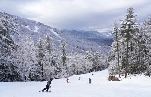 Skiers navigate a snowy slope surrounded by trees and mountains under a cloudy sky.
