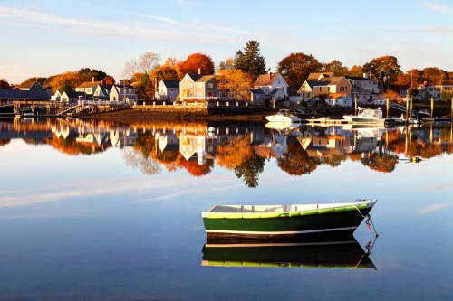 A calm lake reflects colorful autumn trees and houses, with a small green and black boat in the foreground.