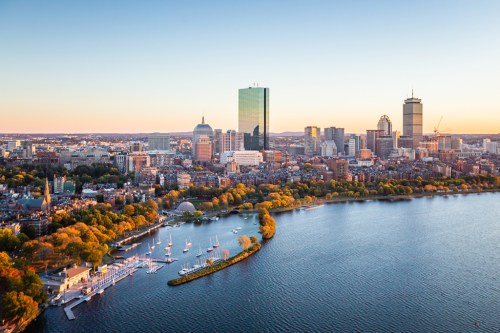 Aerial view of Boston skyline at sunset, featuring skyscrapers and the Charles River with boats and autumn foliage.