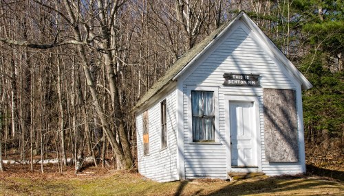 A small, weathered white house with boarded windows, surrounded by bare trees in a rural setting.