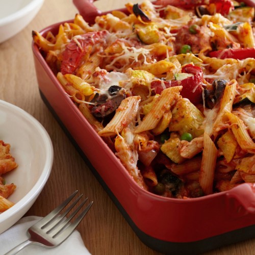 A red casserole dish filled with baked pasta, vegetables, and melted cheese, with a plate and fork nearby.