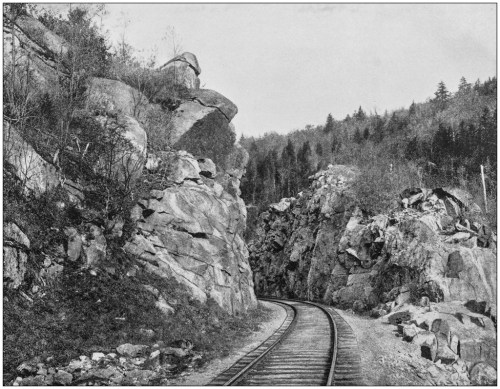A black and white photo of a winding railway track surrounded by rocky cliffs and dense forest.