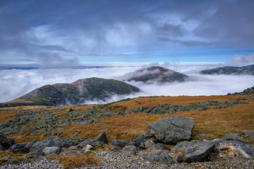 A scenic view of mountains partially covered by clouds, with rocky terrain and patches of grass in the foreground.