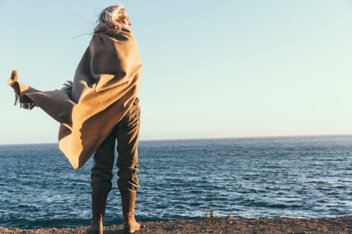 A person with long hair stands by the ocean, wrapped in a blanket, gazing at the horizon during sunset.