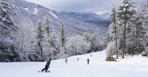 Skiers navigate a snowy slope surrounded by trees and mountains under a cloudy sky.