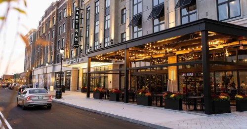 A modern building with outdoor seating, warm lights, and colorful flowers, set in an urban street at dusk.