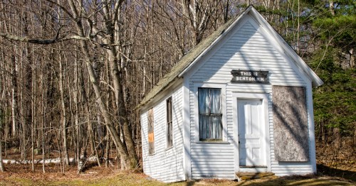 A small, weathered white building with boarded windows, surrounded by bare trees, labeled "THIS IS BENTON, NH."