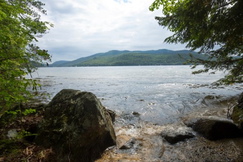 A serene lakeside view with mountains in the background, surrounded by trees and rocky shoreline under a cloudy sky.