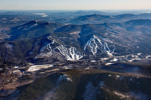 Aerial view of snow-covered mountains with ski trails winding down the slopes, surrounded by dense forests.
