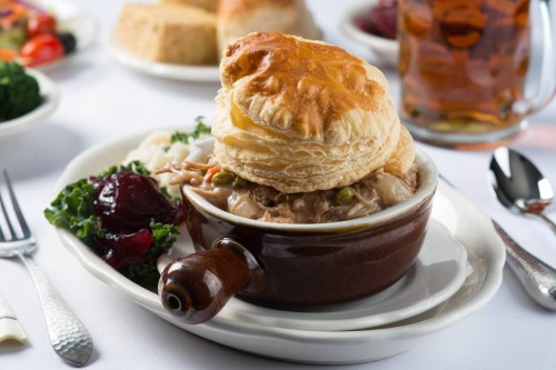 A bowl of chicken pot pie topped with a flaky pastry, served with sides of salad and bread on a white tablecloth.
