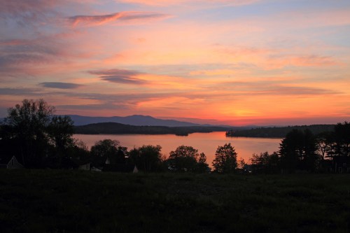 A serene sunset over a lake, with vibrant colors reflecting on the water and silhouettes of trees and mountains in the background.