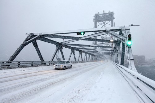 A snowy bridge with a green traffic light and a car driving cautiously on a snow-covered road.