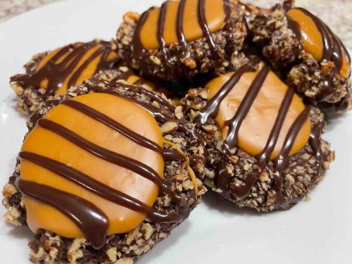 A plate of chocolate-covered cookies topped with caramel and drizzled with chocolate, garnished with nuts.