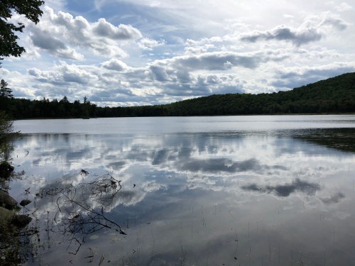 A serene lake reflects clouds and trees, surrounded by lush green hills under a partly cloudy sky.