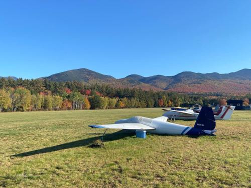 A small airplane parked on a grassy field with colorful autumn trees and mountains in the background under a clear blue sky.
