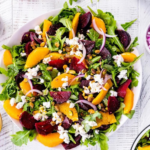 A vibrant salad with arugula, beets, oranges, red onion, goat cheese, and pistachios on a white plate.