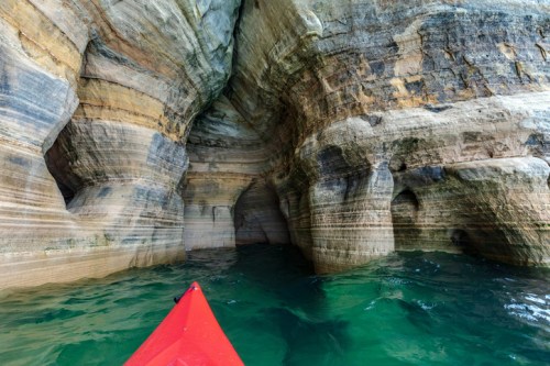 A red kayak glides through a serene cave with layered rock formations and clear turquoise water.