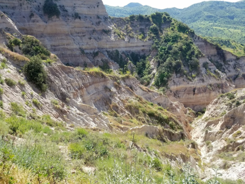 A rugged landscape featuring layered cliffs and greenery, with hills in the background under a clear blue sky.