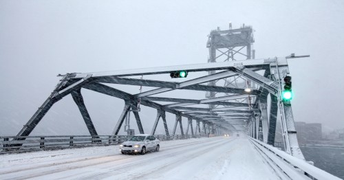 A car drives on a snowy bridge with green traffic lights, surrounded by heavy snowfall and fog.