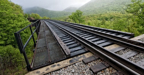 A railway track curves over a bridge, surrounded by lush green trees and misty mountains in the background.