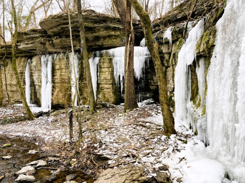 Icicles hang from rocky cliffs in a winter forest, with a stream and snow-covered ground below.