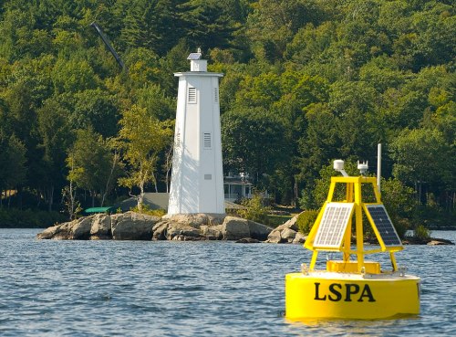 A white lighthouse on a rocky island, with a yellow buoy labeled "LSPA" in the foreground, surrounded by trees and water.