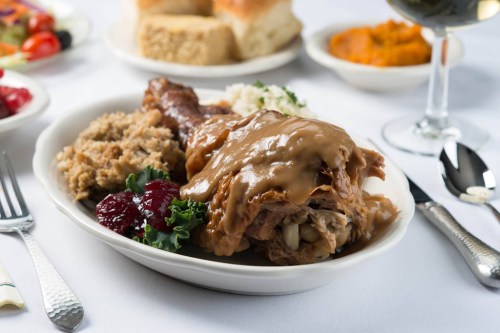 A plate of turkey with gravy, stuffing, cranberry sauce, and sides, served with bread rolls and a glass of wine.