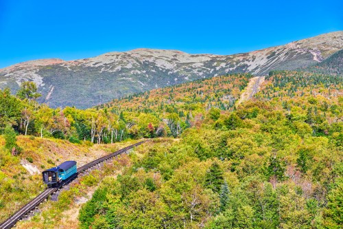 A train travels along a winding track through vibrant autumn foliage with mountains in the background under a clear blue sky.