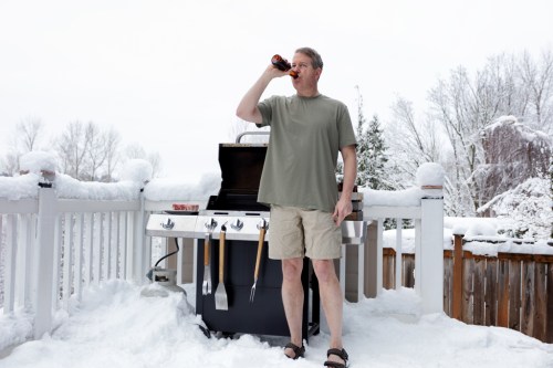 A man in shorts and a t-shirt drinks a beer while standing by a grill on a snowy deck.
