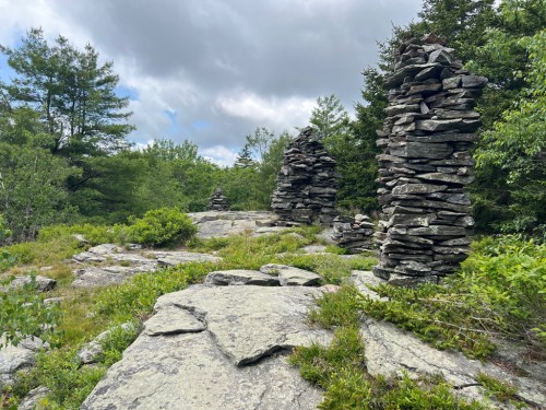 Rock formations stacked among greenery under a cloudy sky, with trees in the background.