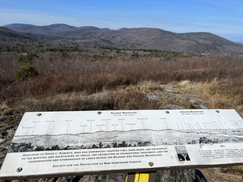 A scenic view of the Belknap and Ossipee Mountains with an informational sign about conservation efforts in the foreground.