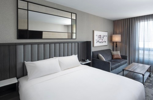 Modern hotel room featuring a bed, sofa, wooden table, and large mirror with natural light from a window.