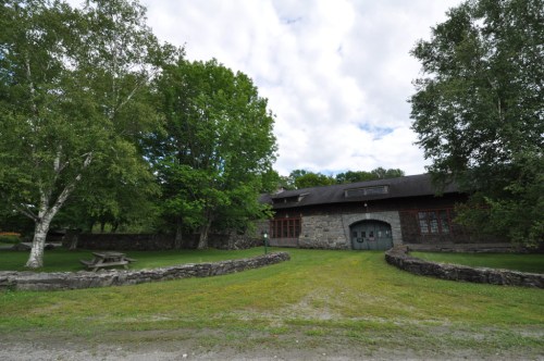 A stone building surrounded by trees and grass, with a picnic table in front and a cloudy sky above.