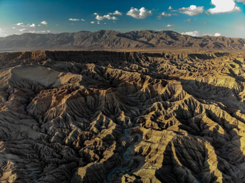 Aerial view of rugged, textured desert landscape with mountains and scattered clouds under a blue sky.