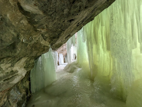 A cave with shimmering green and blue ice formations hanging from the rocky ceiling, reflecting light on the icy ground.