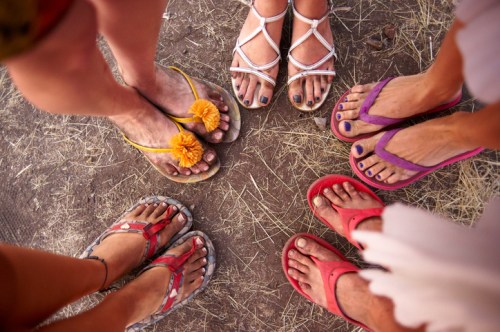 A circle of feet wearing colorful flip-flops on a dirt surface, with some adorned with flowers and painted toenails.