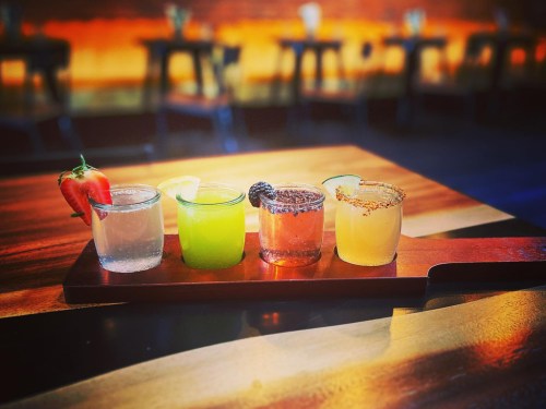 A wooden tray holds five colorful cocktails, each garnished with fruit, set against a blurred restaurant background.