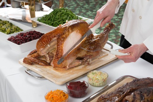 A chef carves a roasted turkey on a wooden board, surrounded by side dishes like green beans and cranberry sauce.